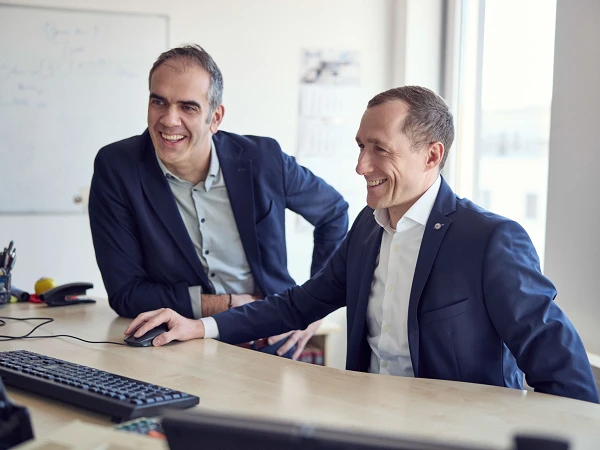 Zwei lächelnde Männer sitzen an einem Schreibtisch, schauen auf einen Computerbildschirm und scheinen eine gemeinsame Aktivität zu genießen. Im Hintergrund sind einige Büromaterialien und ein Fenster mit Tageslicht sichtbar.