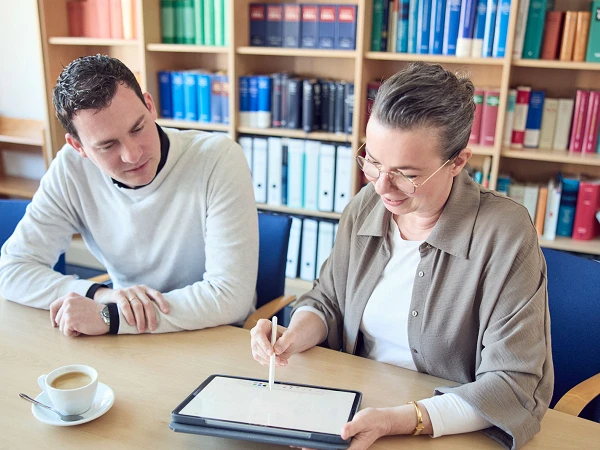 Eine Frau zeigt einem Mann auf einem Tablet eine digitale Präsentation, während sie an einem Tisch in einem Büroumfeld sitzen. Auf dem Tisch stehen eine Tasse Kaffee und ein Glas Wasser. Im Hintergrund sind Regale mit bunten Ordnern zu sehen.