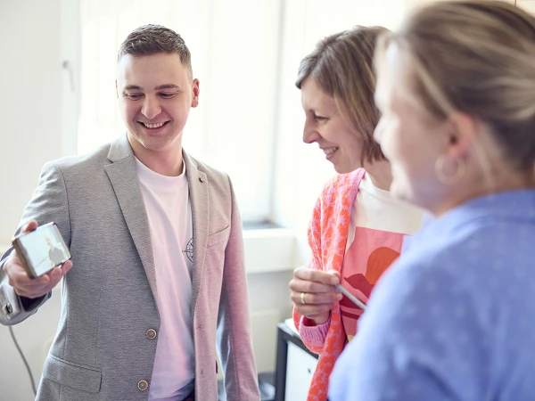 Ein Mann zeigt einer Gruppe von Frauen auf einem Smartphone ein Bild, während sie lächeln und miteinander sprechen. Die Frauen tragen verschiedene farbenfrohe Kleidungsstücke, und der Raum wirkt hell und einladend.