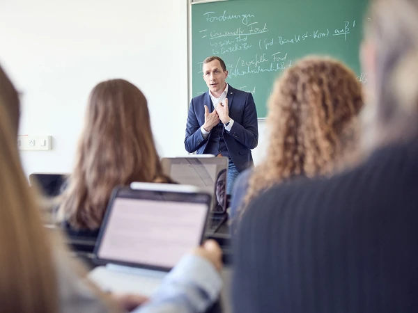 Ein Dozent steht vor einer Tafel und erklärt den Studierenden, die ihm von hinten zugewandt sind, während einige von ihnen auf Laptops arbeiten.