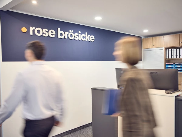 Die Empfangsbereich eines Büros mit dem Schriftzug "rose brösicke" an der Wand, während zwei Personen in Bewegung sind. Am Empfangstresen sind einige Aktenordner sichtbar.