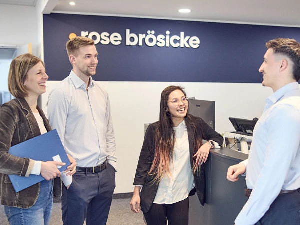 Vier Personen stehen in einem modernen Büro an einem Empfangstresen. Zwei Männer und zwei Frauen unterhalten sich freundlich. Im Hintergrund ist ein Schild mit dem Namen "rose bröschke" zu sehen.