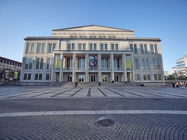 Das Gebäude ist ein großes Theater mit einer eindrucksvollen Fassade, großen Fenstern und einem breiten Treppenaufgang. Vor dem Theater befinden sich Menschen, die sich auf dem Platz bewegen, und es hängen große Banner an der Front des Gebäudes. Der Himmel ist blau und klar.