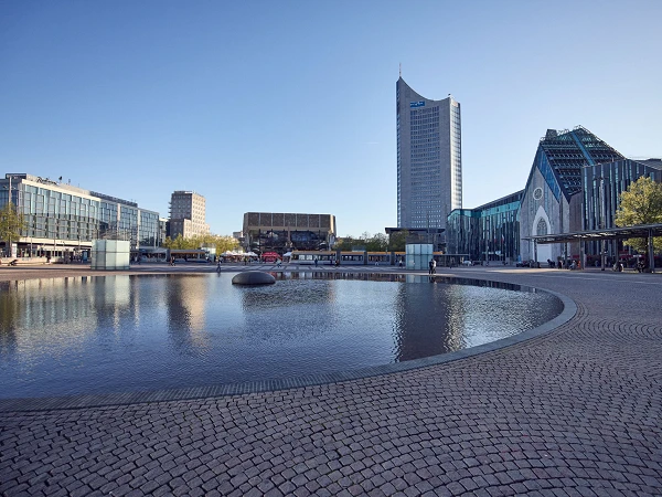 Eine moderne Stadtansicht mit einem großzügigen Platz, auf dem sich ein ruhiger Teich befindet. Im Hintergrund sind mehrere zeitgenössische Gebäude zu sehen, darunter ein markanter Wolkenkratzer und ein architektonisch interessantes Gebäude mit einer Glasfassade. Die Szenerie ist bei klarem Himmel und Sonnenschein festgehalten.