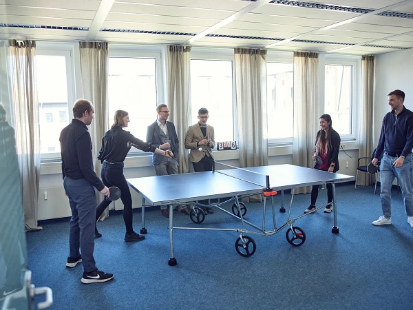 Eine Gruppe von sechs Personen spielt Tischtennis in einem modernen, hellen Raum mit großen Fenstern. Einige Personen sind aktiv am Spielen, während andere zuschauen und anfeuern.