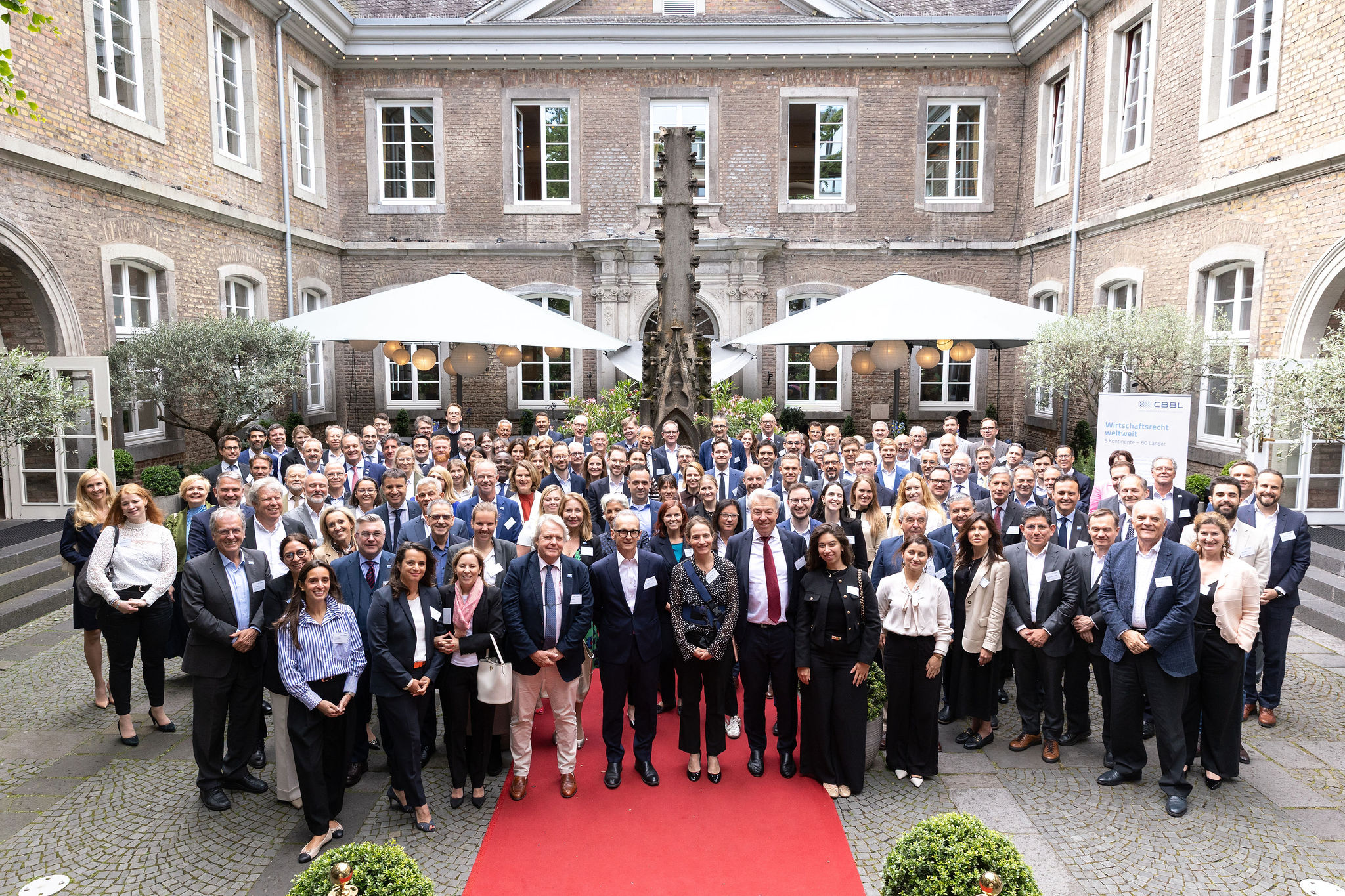 Eine große Gruppe von Menschen steht auf einem roten Teppich in einem Innenhof mit modernen Gebäuden im Hintergrund, umgeben von Pflanzen und Sonnenschirmen, und posiert für ein Gruppenfoto.