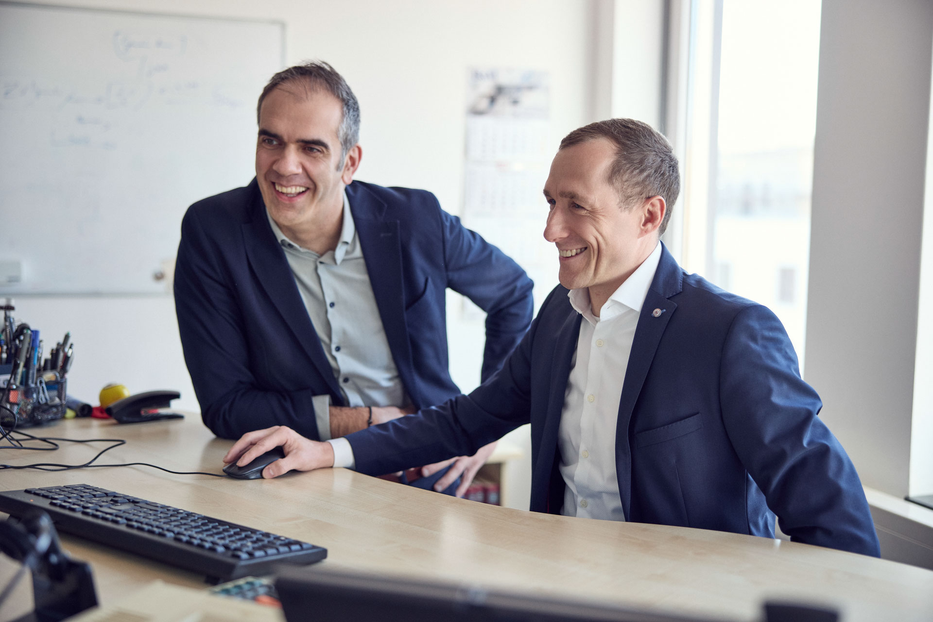 Zwei lächelnde Männer sitzen an einem Schreibtisch, schauen auf einen Computerbildschirm und scheinen eine gemeinsame Aktivität zu genießen. Im Hintergrund sind einige Büromaterialien und ein Fenster mit Tageslicht sichtbar.