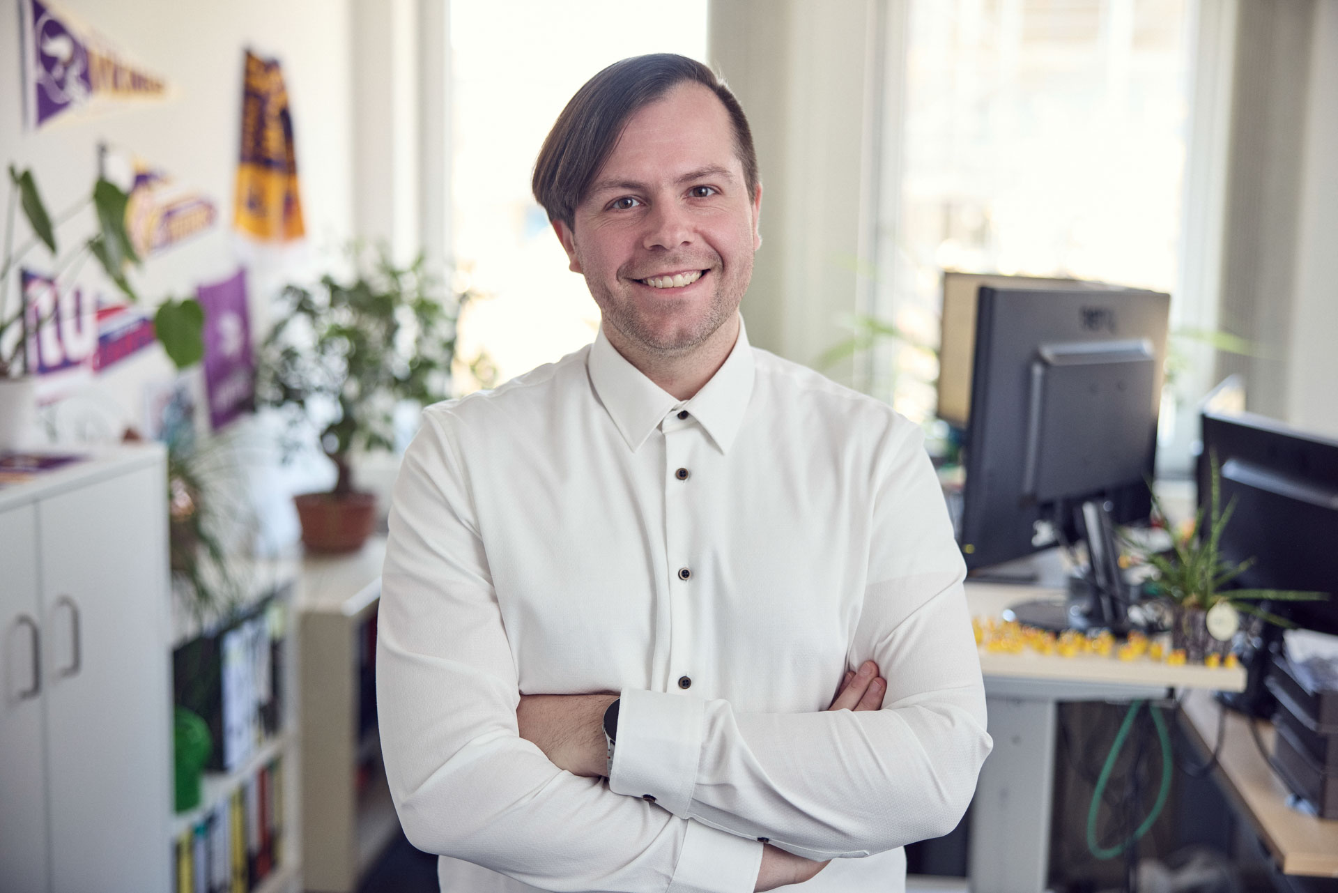 Ein lächelnder Mann steht mit verschränkten Armen in einem hellen Büro, umgeben von Pflanzen und Büromöbeln. Hinter ihm sind Regale und ein Computer sichtbar.