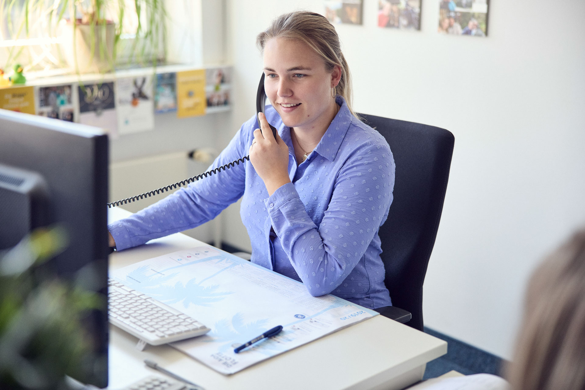 Eine Frau mit blonden Haaren sitzt an einem Schreibtisch, spricht am Telefon und schaut auf einen Computerbildschirm, während sie Notizen auf einem Schreibblock macht.
