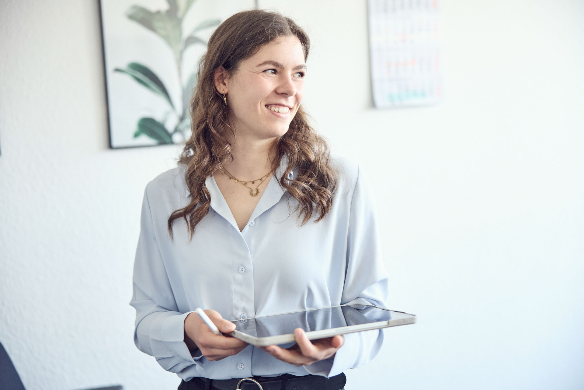 Eine lächelnde Frau mit langen, lockigen Haaren trägt ein hellblaues Hemd und hält ein Tablet in der Hand, während sie in einem modernen Raum steht, der mit Pflanzen und einem Kalender an der Wand dekoriert ist.