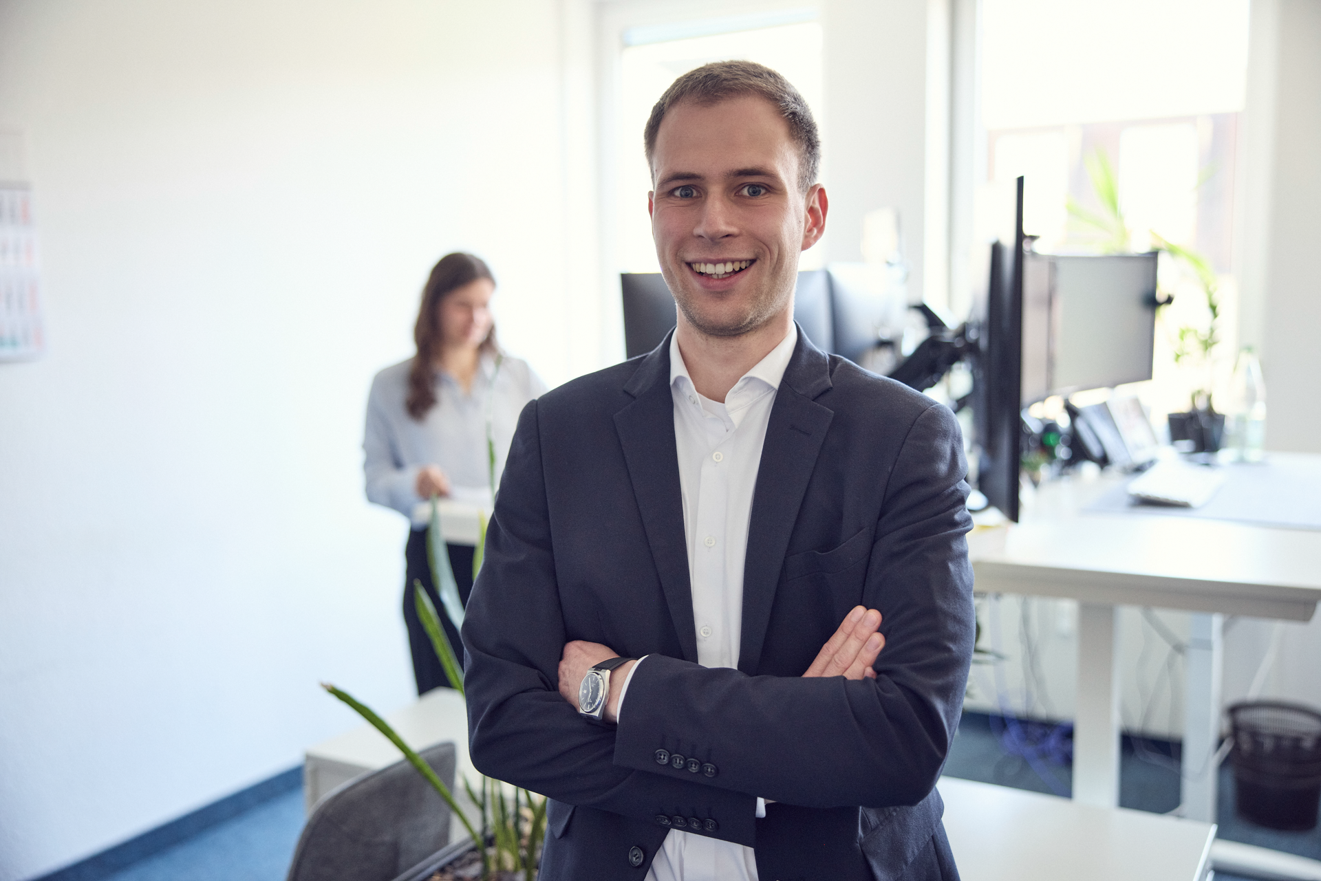 Ein lächelnder Mann in einem Anzug steht mit verschränkten Armen in einem modernen Büroraum, während im Hintergrund eine Frau an einem Schreibtisch arbeitet. Pflanzen und Computer sind im Raum sichtbar.