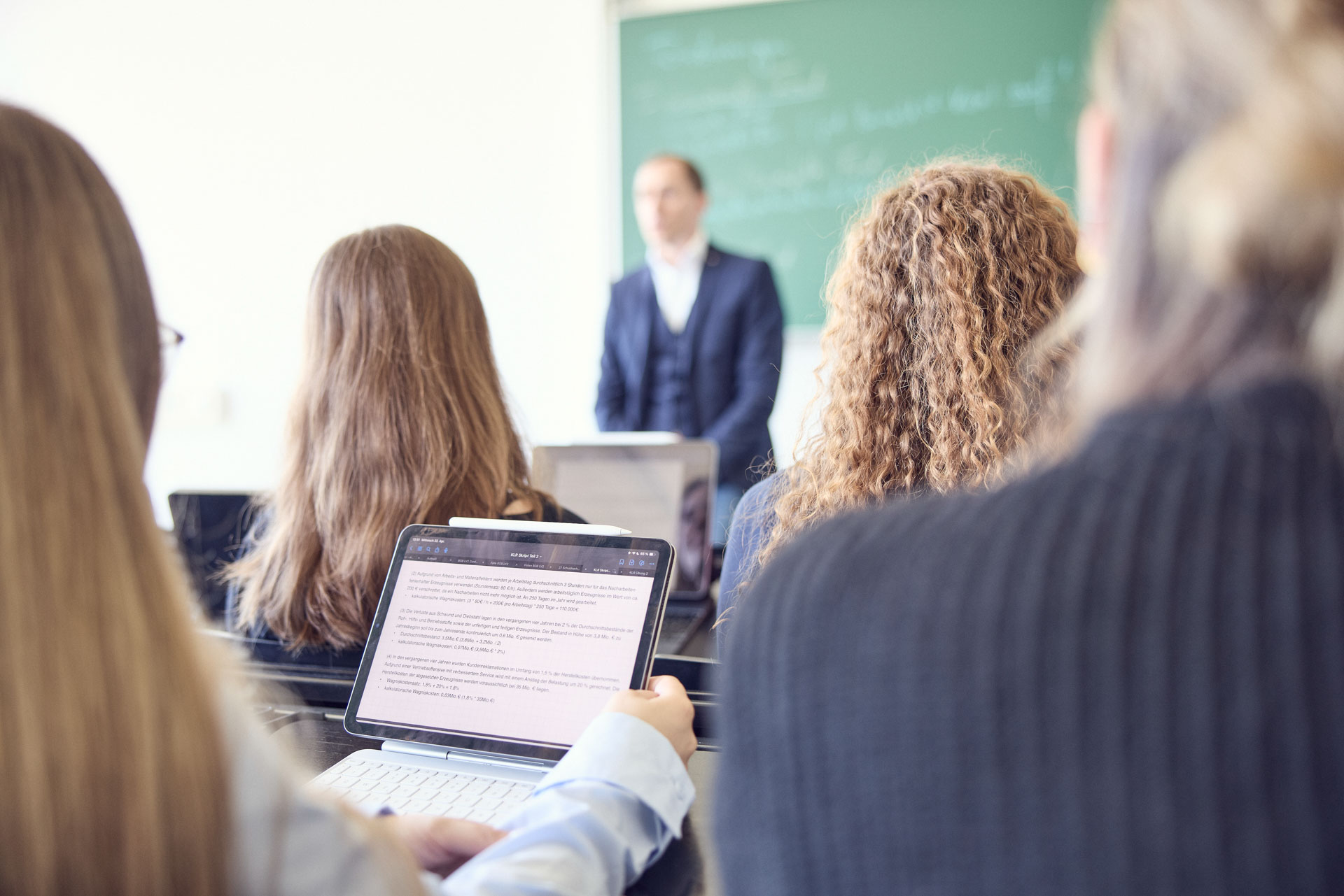 Eine Gruppe von Studierenden sitzt in einem Seminarraum und blickt auf einen Dozenten, der vor einer grünen Tafel spricht. Einige Studierende verwenden Laptops, während sie dem Unterricht folgen.