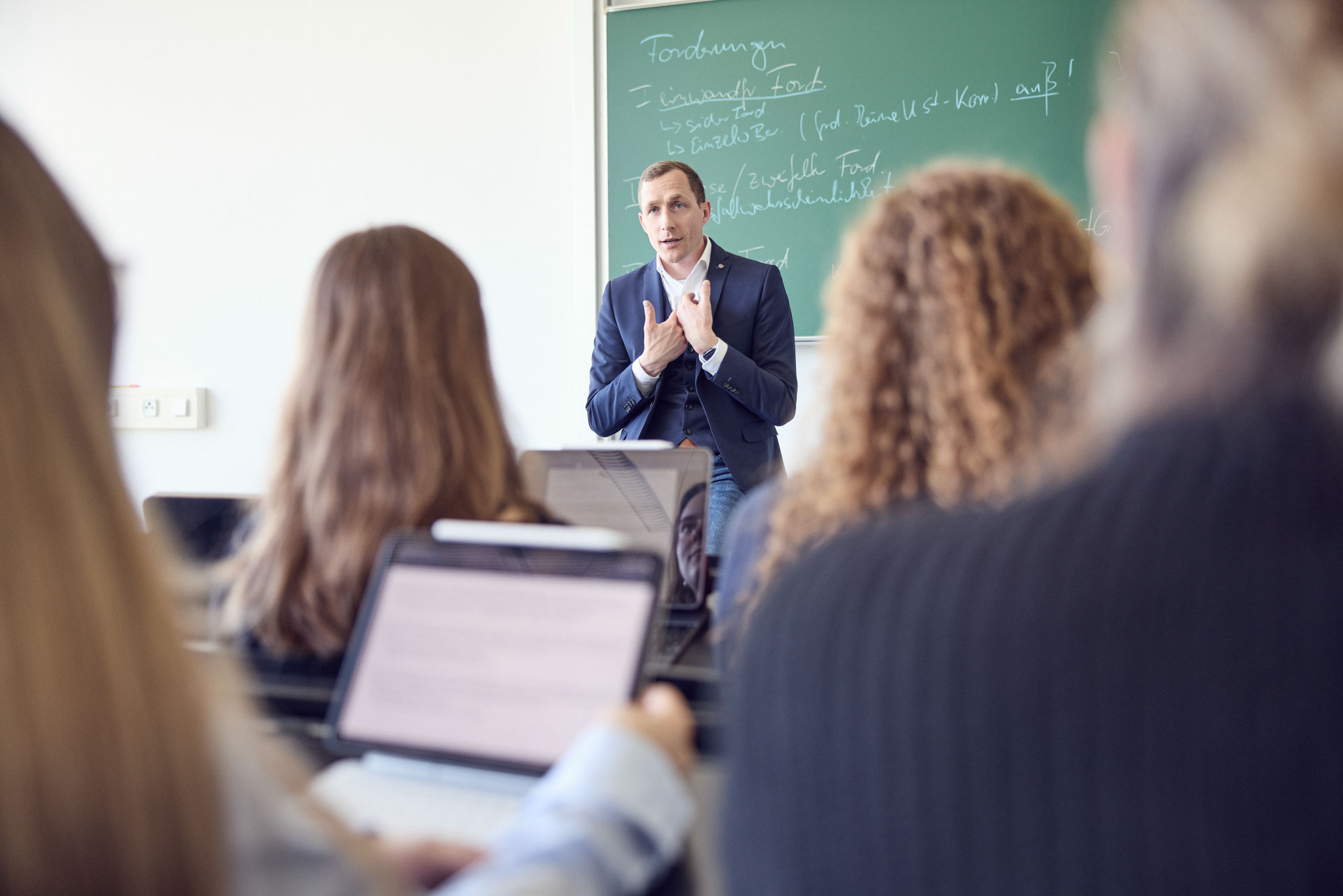 Ein Dozent steht vor einer Tafel und erklärt den Studierenden, die ihm von hinten zugewandt sind, während einige von ihnen auf Laptops arbeiten.