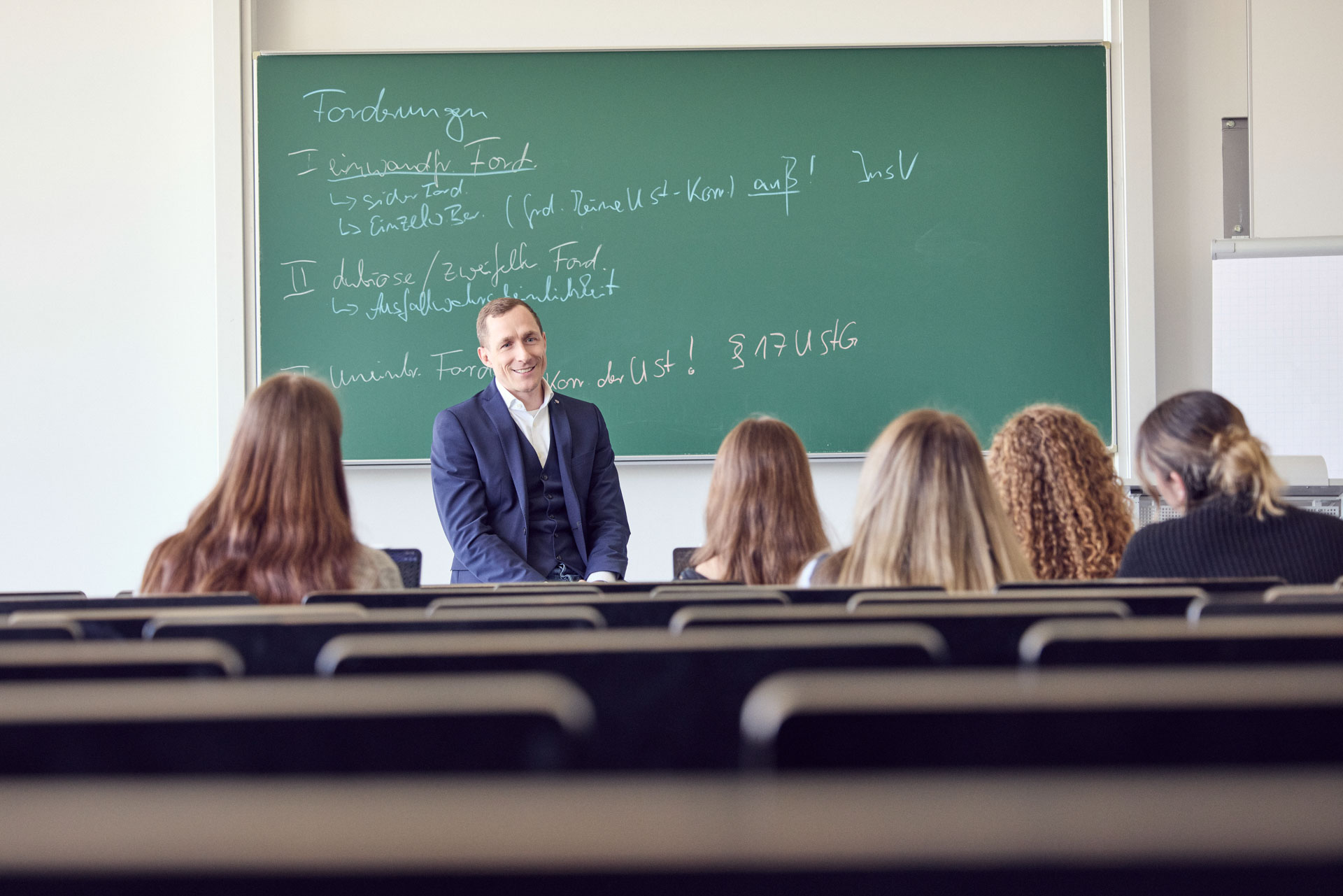 Ein Lehrer sitzt vor einer Gruppe von Studierenden in einem Seminarraum und erklärt Inhalte, während an einer Tafel mathematische Formeln und Notizen geschrieben sind.