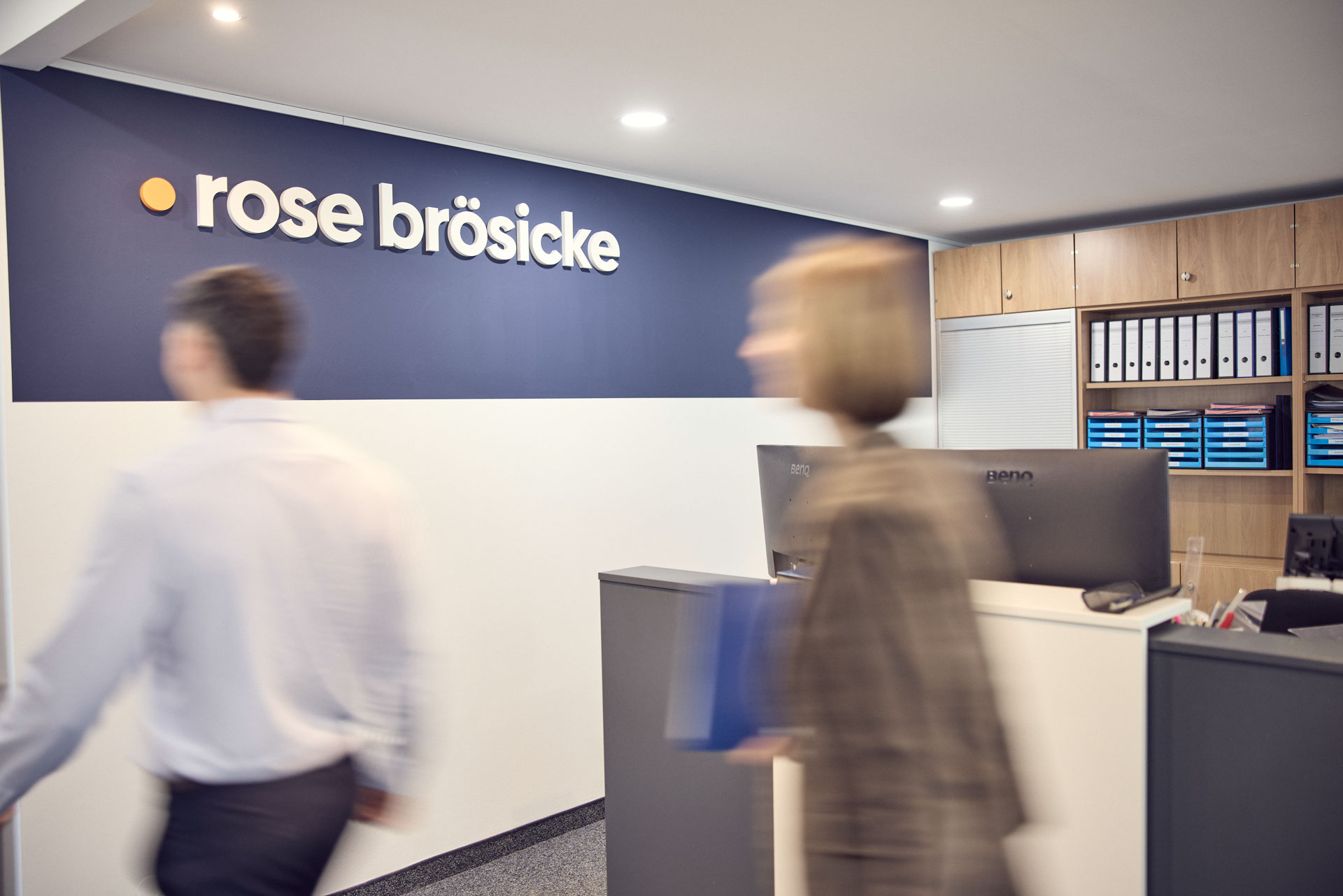 Die Empfangsbereich eines Büros mit dem Schriftzug "rose brösicke" an der Wand, während zwei Personen in Bewegung sind. Am Empfangstresen sind einige Aktenordner sichtbar.