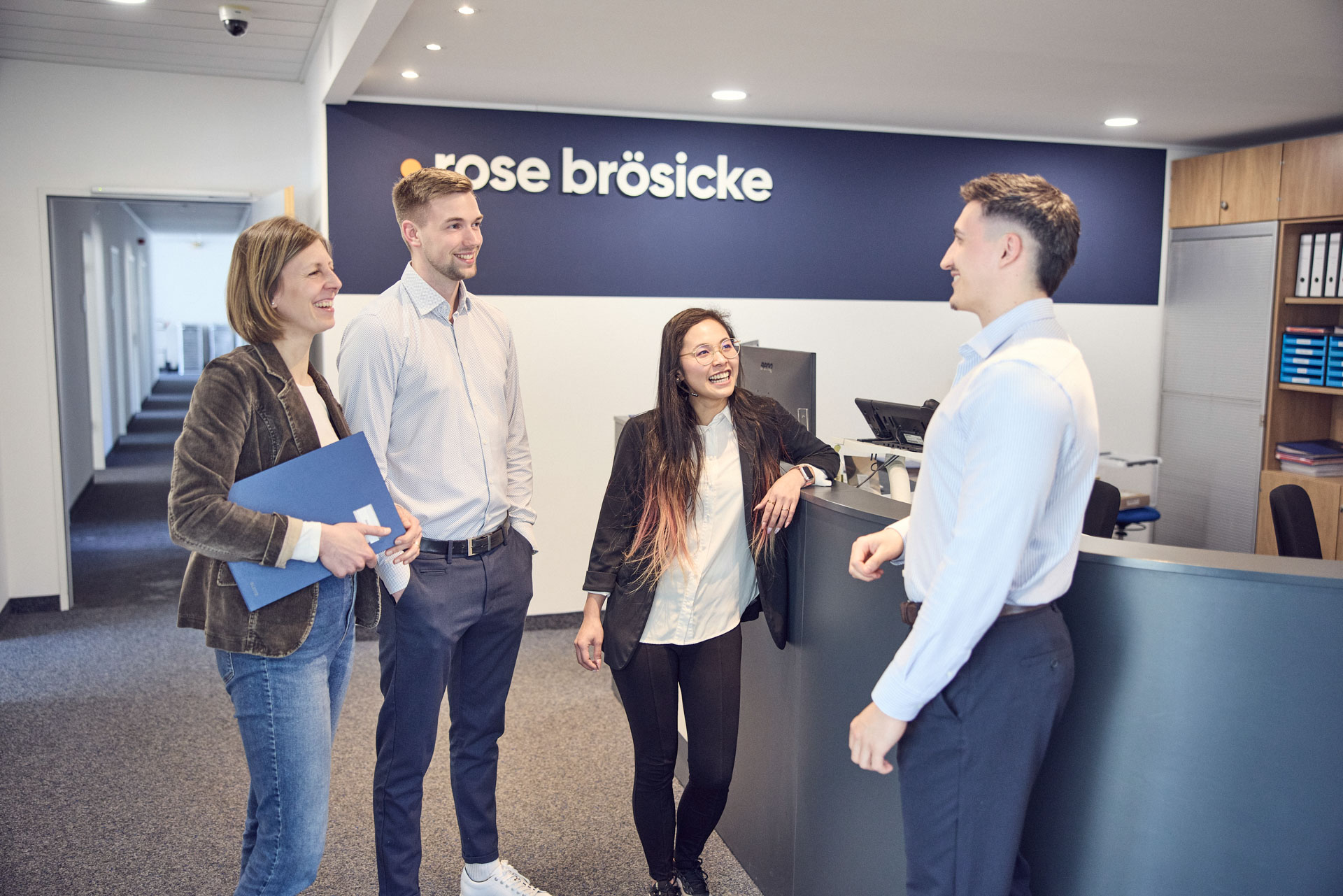 Vier Personen stehen in einem modernen Büro an einem Empfangstresen. Zwei Männer und zwei Frauen unterhalten sich freundlich. Im Hintergrund ist ein Schild mit dem Namen "rose bröschke" zu sehen.