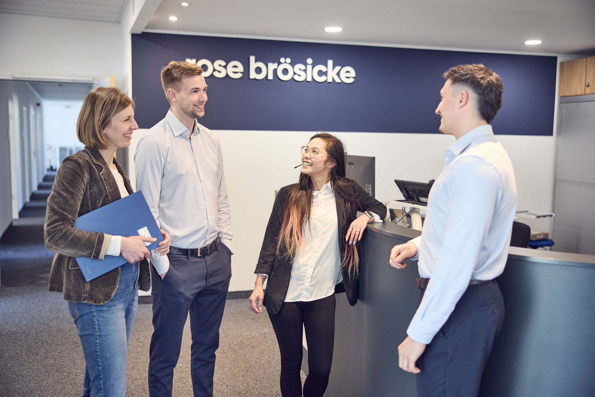 Eine Gruppe von vier Personen steht an einem Empfangstresen und unterhält sich freundlich; eine Frau hält eine Mappe, während eine andere mit einem Headset lächelt. Im Hintergrund ist ein Schild mit der Aufschrift "rose brösicke" sichtbar.