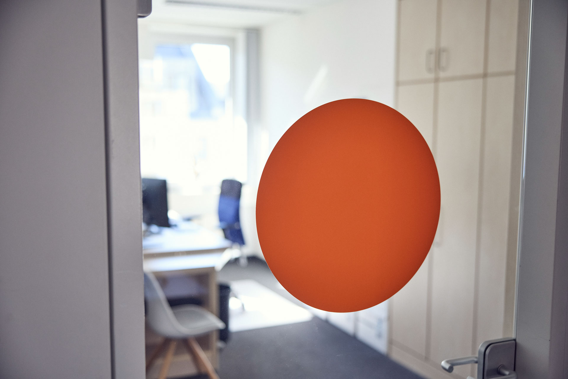 Ein orangefarbener, runder Aufkleber ist auf einer Glastür angebracht, hinter der ein hell erleuchteter Büroraum mit Schreibtisch und Stühlen zu sehen ist.