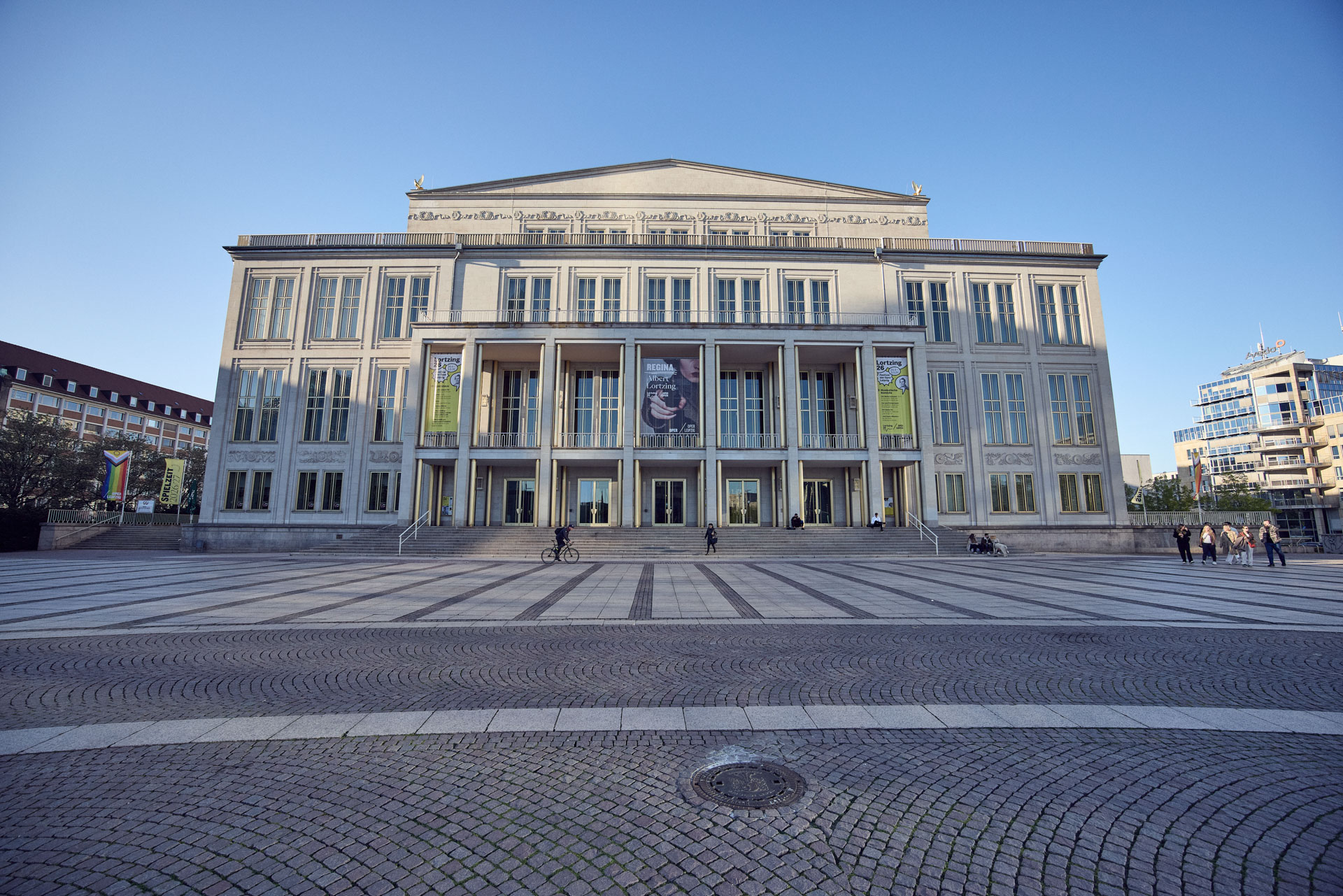 Das Gebäude ist ein großes Theater mit einer eindrucksvollen Fassade, großen Fenstern und einem breiten Treppenaufgang. Vor dem Theater befinden sich Menschen, die sich auf dem Platz bewegen, und es hängen große Banner an der Front des Gebäudes. Der Himmel ist blau und klar.