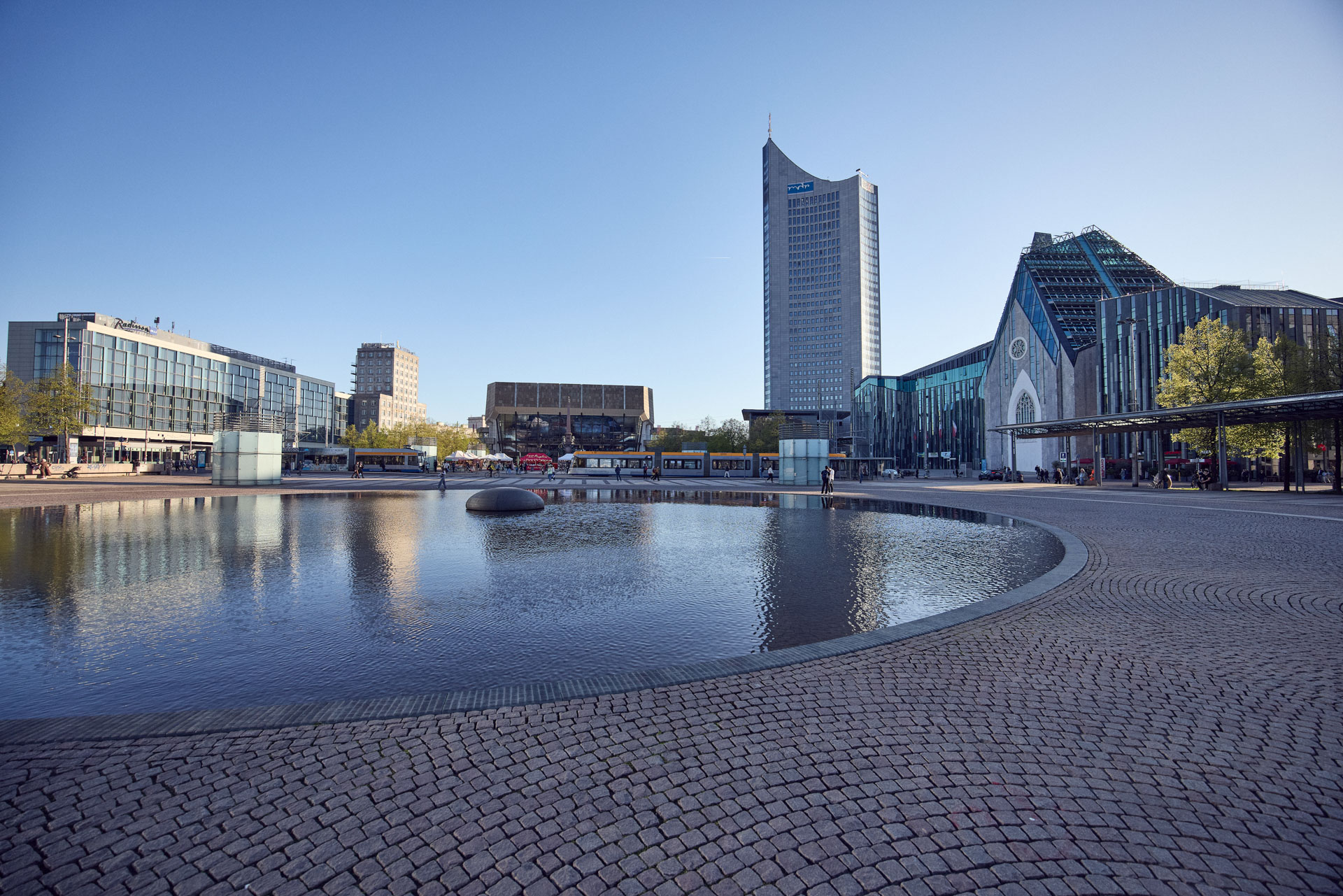 Eine moderne Stadtansicht mit einem großzügigen Platz, auf dem sich ein ruhiger Teich befindet. Im Hintergrund sind mehrere zeitgenössische Gebäude zu sehen, darunter ein markanter Wolkenkratzer und ein architektonisch interessantes Gebäude mit einer Glasfassade. Die Szenerie ist bei klarem Himmel und Sonnenschein festgehalten.
