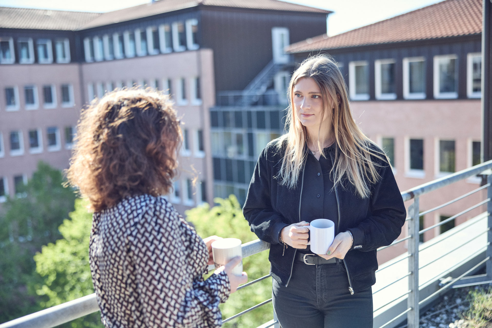 Zwei Frauen stehen auf einem Balkon und unterhalten sich, während sie Tassen in der Hand halten. Im Hintergrund sind mehrstöckige Gebäude und Bäume zu sehen.