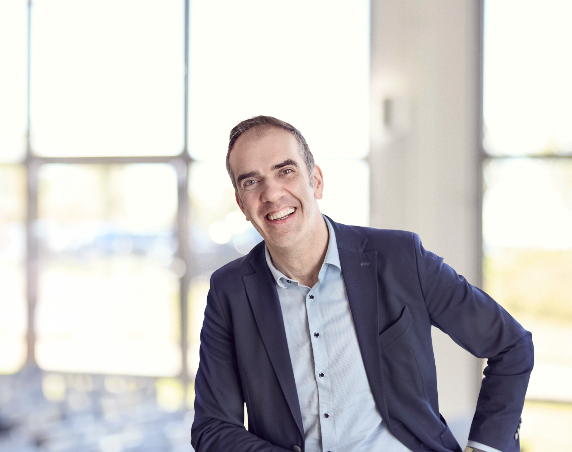Ein Mann in einem blazer sitzt lächelnd in einem hellen Raum mit großen Fenstern im Hintergrund.