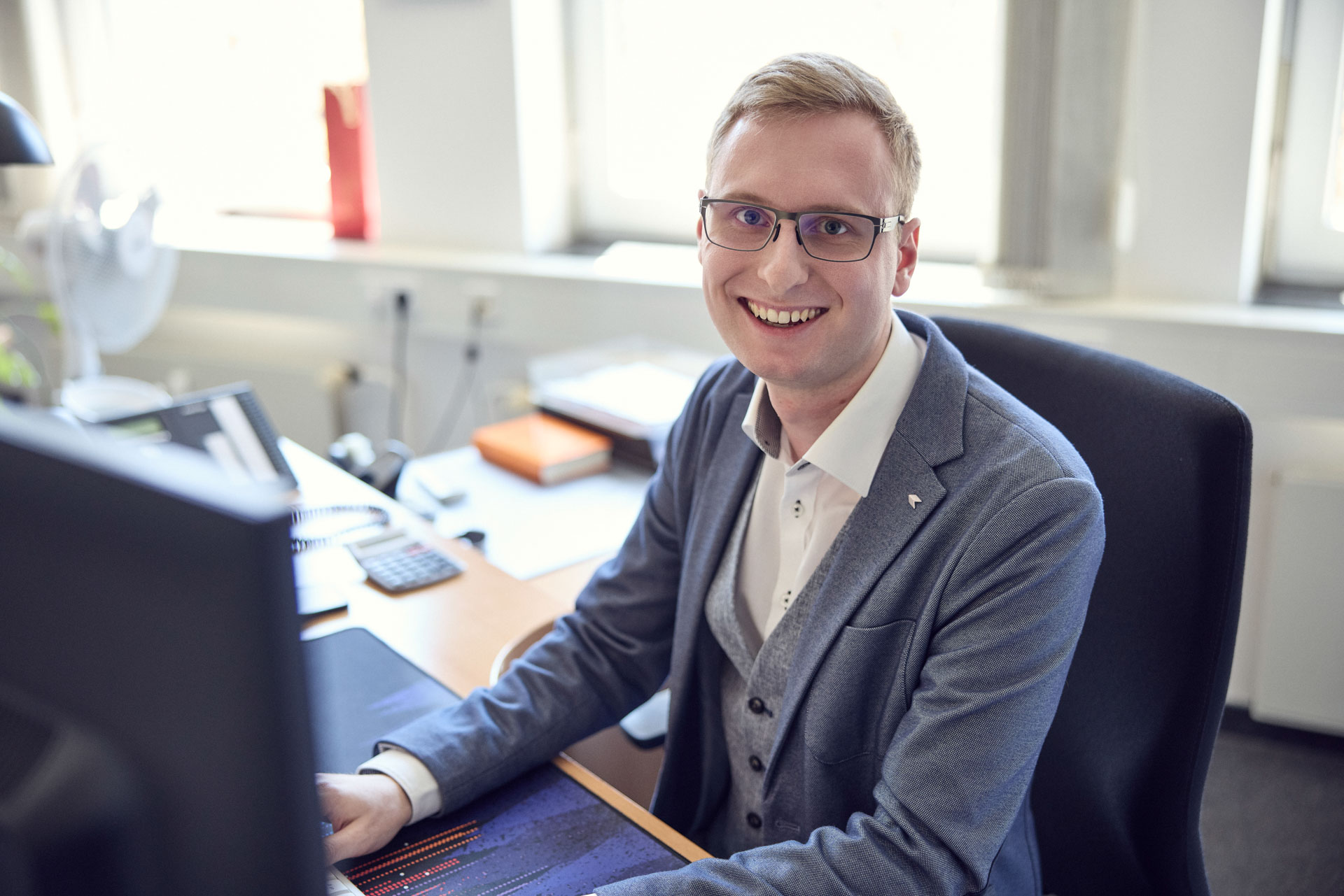 Ein junger Mann mit Brille und blonder, kurzer Haarfrisur sitzt an einem Schreibtisch und lächelt in die Kamera. Er trägt einen grauen Blazer über einem weißen Hemd und ist umgeben von Büroausstattung, darunter ein Computer und Schreibmaterialien.