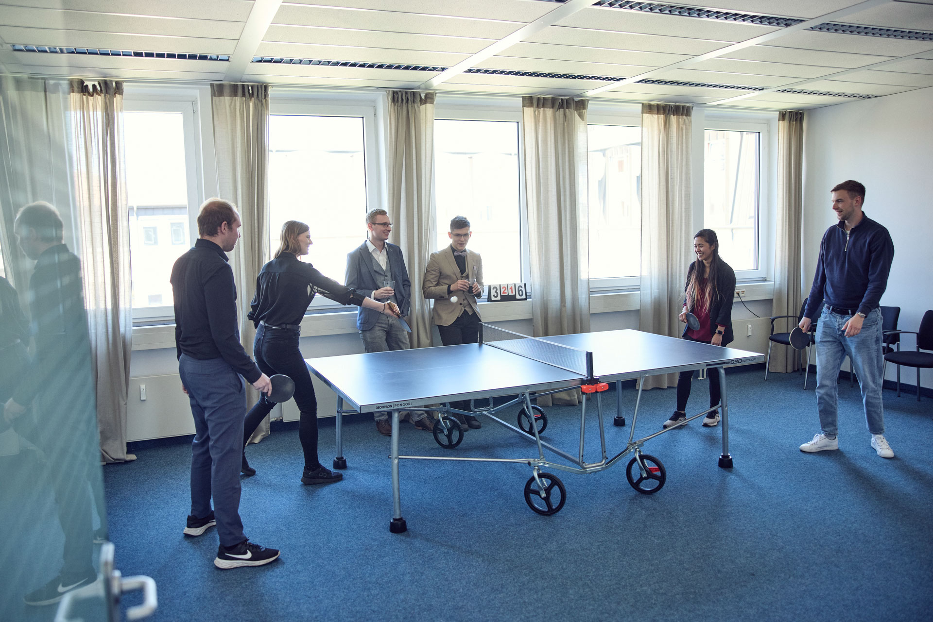 Eine Gruppe von sechs Personen spielt Tischtennis in einem modernen, hellen Raum mit großen Fenstern. Einige Personen sind aktiv am Spielen, während andere zuschauen und anfeuern.