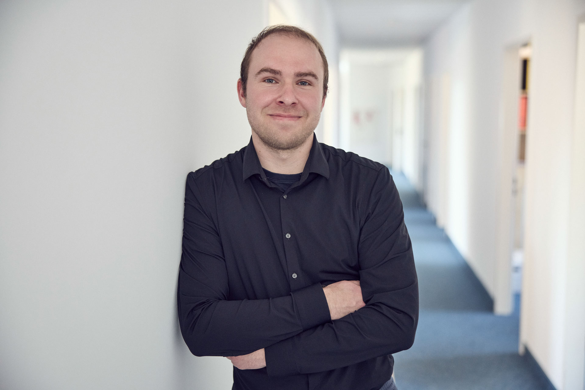Ein Mann mit kurzen Haaren und einem schwarzen Hemd steht lächelnd in einem hellen Flur, die Arme vor der Brust verschränkt, während er an einer Wand lehnt.