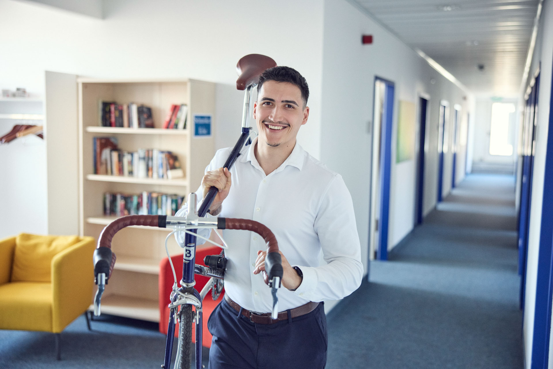 Ein junger Mann mit Schnurrbart trägt ein Fahrrad in einem modernen, hellen Flur, während er lächelt. Im Hintergrund sind Bücherregale und eine gelbe Couch zu sehen.