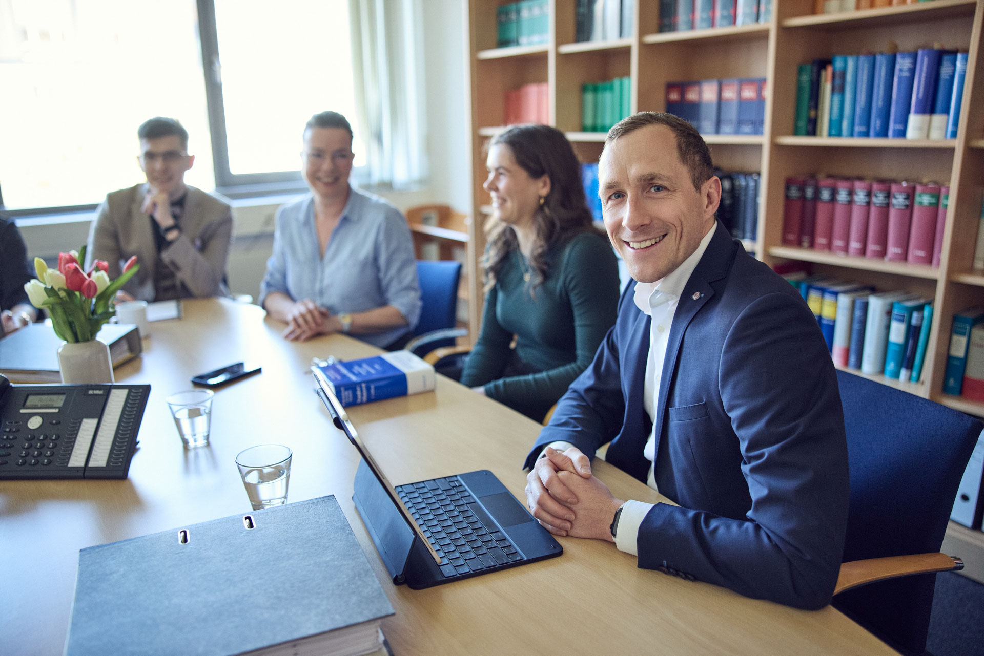 Eine Gruppe von vier Personen sitzt an einem Konferenztisch in einem Büro, lächelnd und sich unterhaltend. Vor ihnen liegt ein Stapel Bücher, und auf dem Tisch befindet sich ein Laptop sowie eine Telefonanlage. Eine Person in blauer Kleidung schaut der Kamera freundlich entgegen. Im Hintergrund sind Regale mit bunten Buchrücken zu sehen.