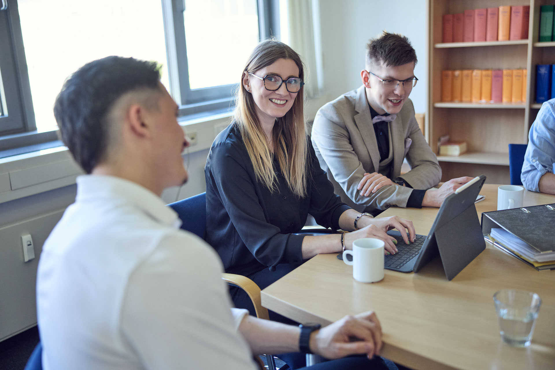 Eine Gruppe von vier Personen in einem Besprechungsraum, die an einem Tisch sitzen. Eine Frau mit langen, blonden Haaren und Brille lächelt, während sie auf ein Tablet tippt. Die anderen Personen, zwei Männer und eine weitere Frau, hören aufmerksam zu. Auf dem Tisch stehen Tassen und einige Bücher sind im Hintergrund sichtbar.