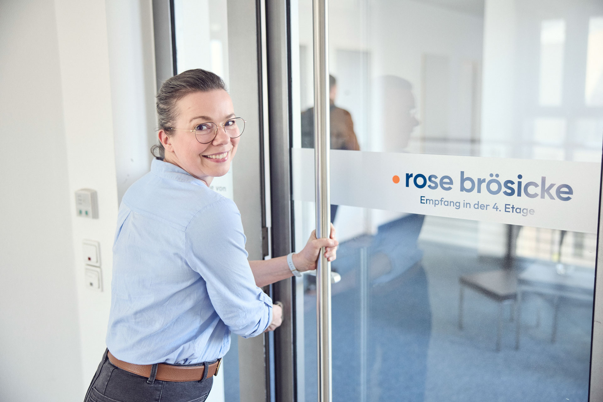 Eine Frau mit Brille lächelt, während sie eine Glastür mit der Aufschrift „rose brösicke Empfang in der 4. Etage“ öffnet. Im Hintergrund ist ein weiterer Raum zu sehen.